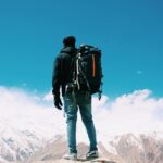 A lone hiker with a backpack surveys snow-capped mountains under a clear blue sky.