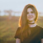 Portrait of a smiling young woman outdoors in a sunlit field, wearing a black choker.